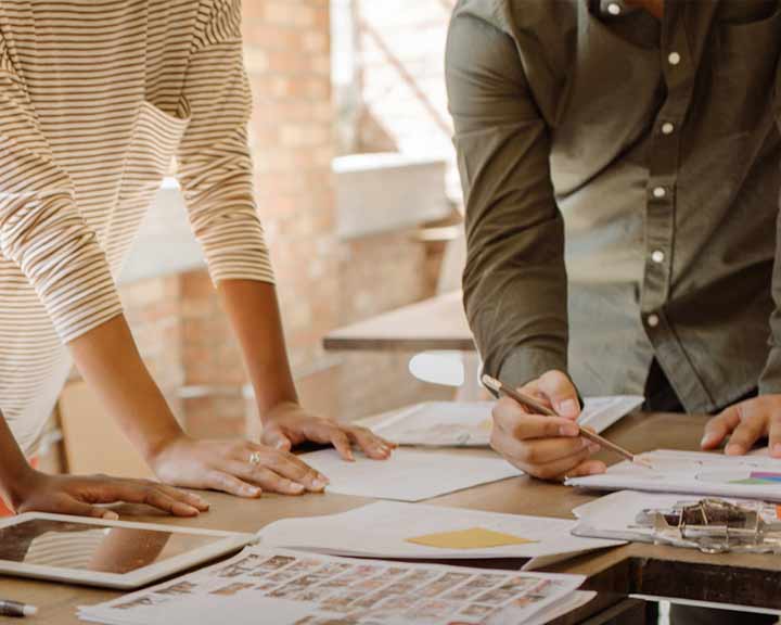 Deux personnes sont assises autour d'une table, consultant des documents et une tablette. L'une d'elles tient un stylo, semblant prendre des notes ou discuter des documents.