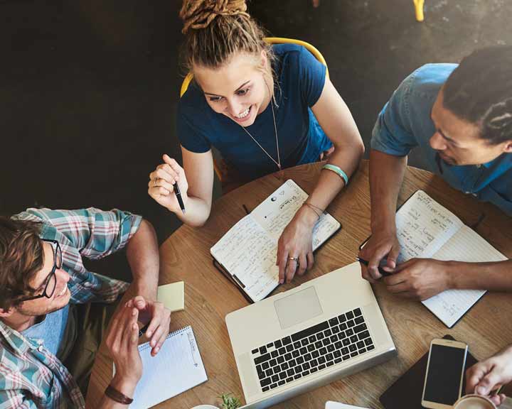 Trois personnes sont assises autour d'une table ronde, avec un ordinateur portable, des cahiers et un téléphone portable. Ils semblent travailler ensemble et discuter.