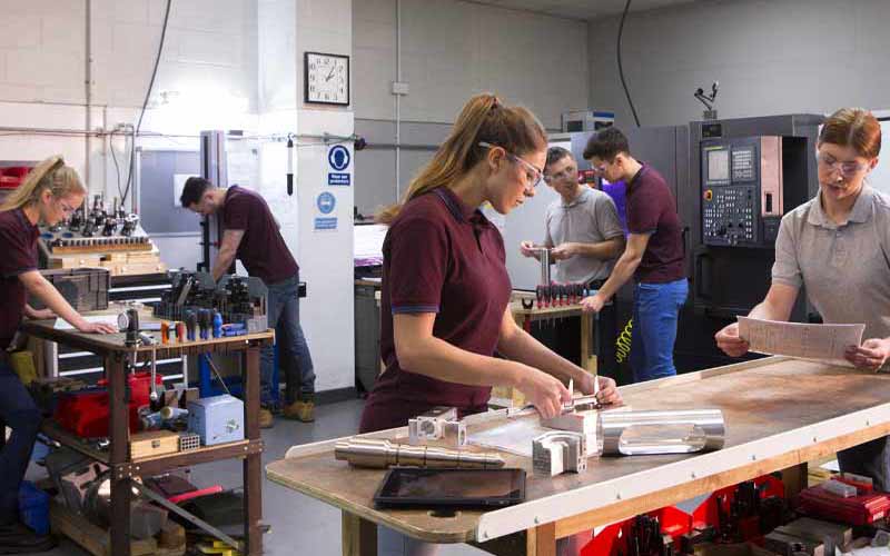 Étudiants.e.s, tous occupés à travailler dans un atelier. Les mesures et le travail du métal sont minutieux. Les jeunes étudiants.e.s suivent les conseils d'adultes plus expérimentés. Établis, outils, équipements et machines remplissent la pièce.