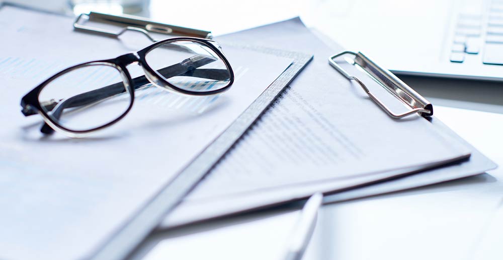 Une paire de lunettes à monture noire repose sur des papiers et des porte-documents, un ordinateur portable étant visible en arrière-plan. La scène évoque un environnement de travail ou d'études.