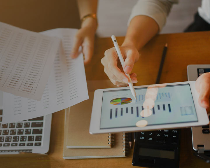 Une personne consulte des documents financiers à l'aide d'un stylet sur une tablette affichant des graphiques. L'image représente un environnement de travail avec un ordinateur portable et des documents.