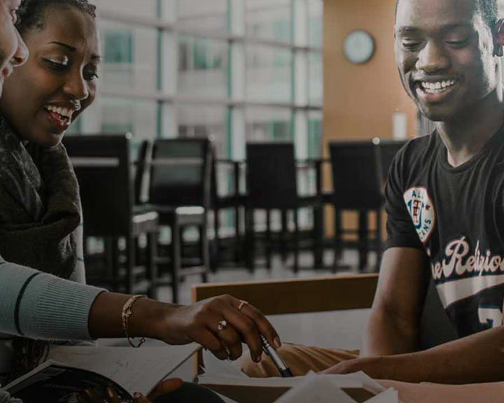 Trois jeunes adultes noirs sont réunis autour d'une table, souriants et absorbés par une discussion. L'un d'eux désigne des papiers posés sur la table, laissant supposer qu'ils travaillent sur un projet ou étudient ensemble.