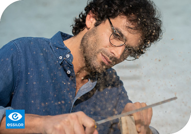 Un artisan concentré, portant des lunettes rondes, travaille méticuleusement sur un morceau de bois, la sciure de bois pulvérisant autour de lui, créant une atmosphère dynamique et légèrement granuleuse.
