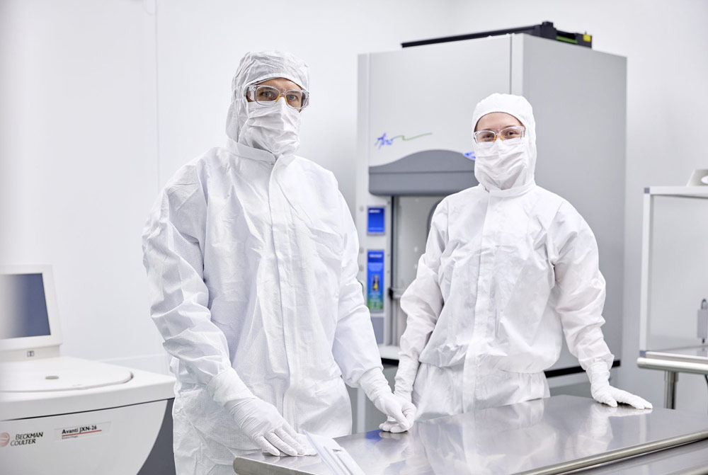 Deux personnes en tenue stérile complète se tiennent dans un laboratoire propre. Elles portent des équipements de protection et travaillent dans un environnement contrôlé, probablement lié à la recherche biomédicale.