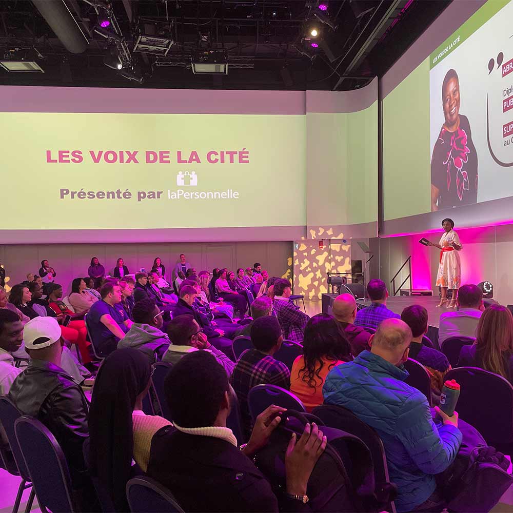 L'image représente une foule assise lors d'une présentation intitulée "Les Voix de la Cité", où une femme est sur scène en train de parler.