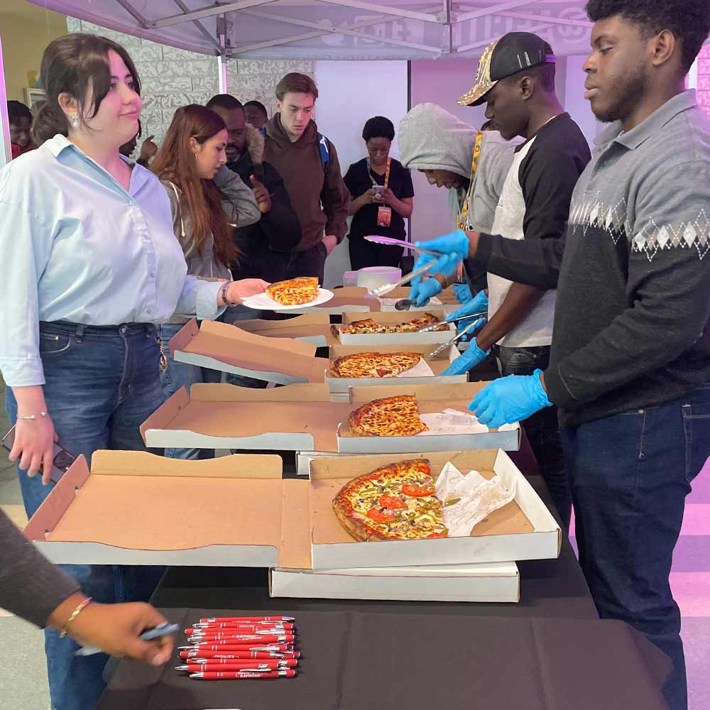 L'image montre une ligne de personnes qui se font servir des parts de pizza dans des boîtes à pizza ouvertes lors d'un événement ou d'un rassemblement social. Devant les boîtes à pizza, des gens attendent d'être servis, tandis que des parts sont distribuées avec des pinces par des serveurs portant des gants en plastique bleus.