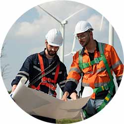 Deux ingénieurs, équipés d'équipements de sécurité, examinent ensemble des plans dans un parc éolien par une journée ensoleillée, avec des éoliennes en arrière-plan, reflétant le travail d'équipe et l'attention portée à l'environnement.