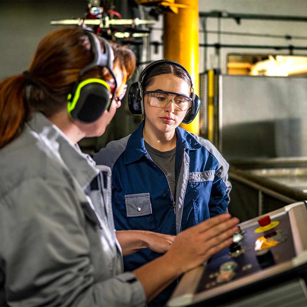 A mature female CNC operator in protective ear gear offers guidance to a younger apprentice using water jet cutting technology within an industrial setting.