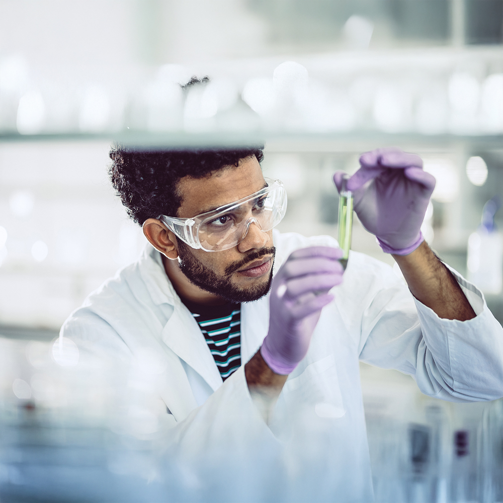 Jeune scientifique examinant un tube à essai contenant un liquide vert dans un laboratoire, portant blouse, gants et lunettes de protection, illustrant concentration et rigueur scientifique.