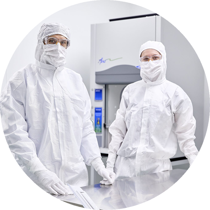 Deux personnes en salle blanche, entièrement vêtues de combinaisons de protection blanches avec masques, gants et lunettes, travaillant derrière un plan en inox devant un équipement scientifique sophistiqué, dans un environnement stérile et lumineux.