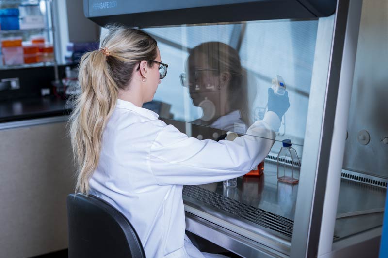 Scientifique féminine travaillant dans un laboratoire, manipulant une pipette derrière une enceinte en verre, illustrant rigueur et précision dans un environnement stérile et organisé.