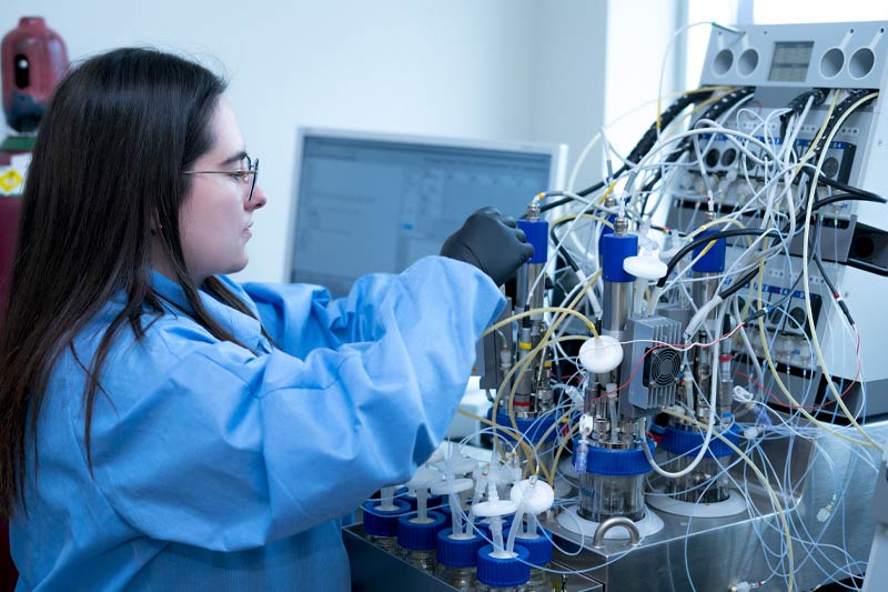 Jeune scientifique en laboratoire, portant une blouse bleue et des gants noirs, concentrée sur un complexe dispositif expérimental composé de flacons, tubes et câblage, dans un environnement stérile et technique.