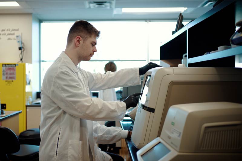 Laboratoire avec un jeune scientifique en blouse et gants noirs utilisant un équipement complexe, autres appareils et collègue flou en arrière-plan.
