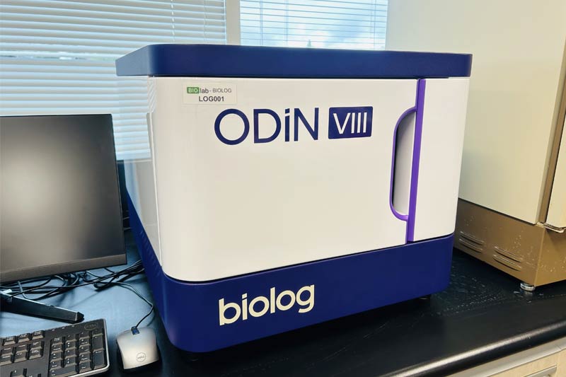 Laboratoire avec la machine « ODiN VIII » de Biolog, accompagnée d’un ordinateur pour son contrôle, câbles visibles, et lumière naturelle entrant par une fenêtre en arrière-plan.