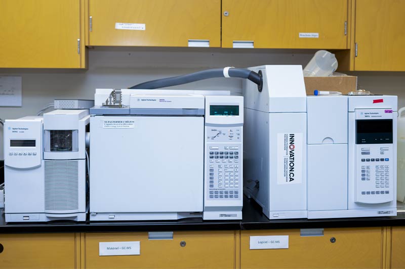 Laboratoire avec plusieurs appareils analytiques blancs, panneaux de contrôle visibles, marques comme Agilent, sur un comptoir noir, avec armoires jaunes et fournitures de laboratoire en arrière-plan.