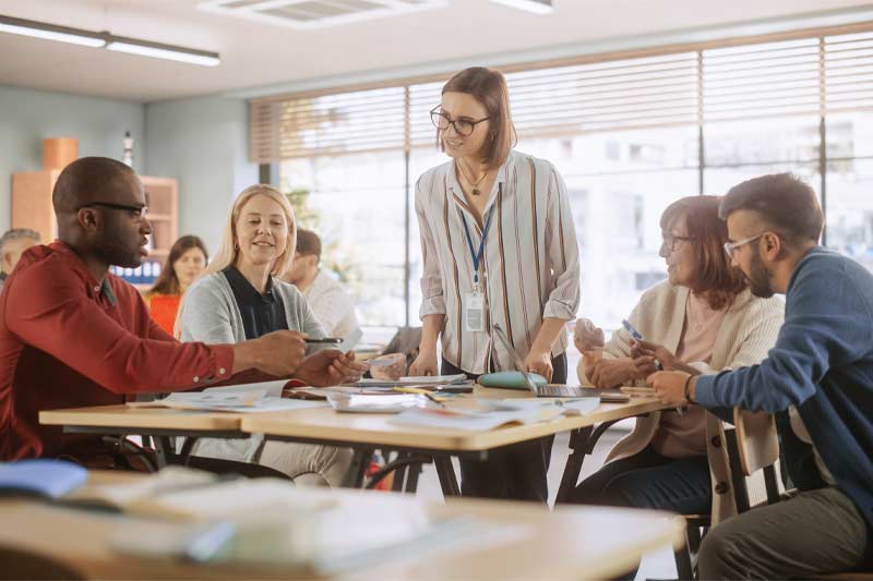 Une salle de classe lumineuse et interactive où une enseignante souriante anime une discussion avec des élèves engagés, de diverses origines, travaillant en groupe.