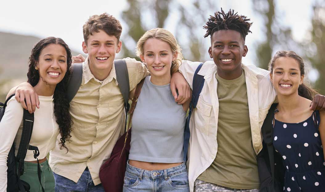 Cinq jeunes amis, aux profils variés, sacs à dos sur le dos, sourient joyeusement en posant ensemble extérieur. Bras dessus bras dessous, ils ont l'air prêts pour l'aventure.