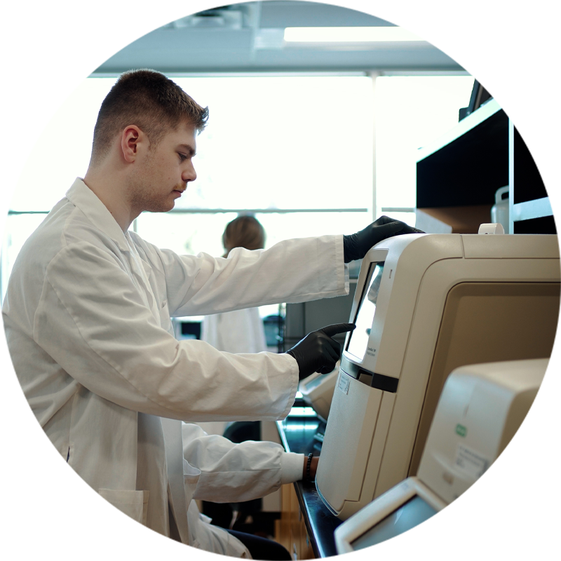 Jeune homme en laboratoire manipulant un équipement scientifique sophistiqué, portant une blouse blanche et des gants, dans un environnement lumineux et professionnel.