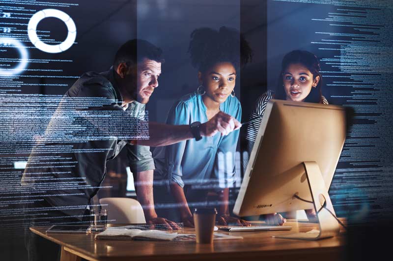 Trois personnes collaborent autour d’un écran d’ordinateur dans un bureau sombre, avec des éléments visuels suggérant l’analyse de données et la technologie.