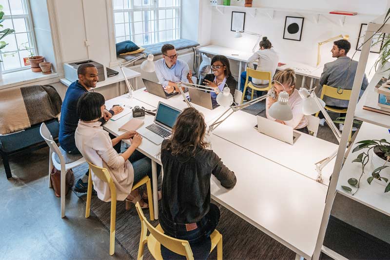 Plusieurs personnes travaillent et échangent autour de grands bureaux blancs, utilisant des ordinateurs portables dans un espace de travail moderne et collaboratif.