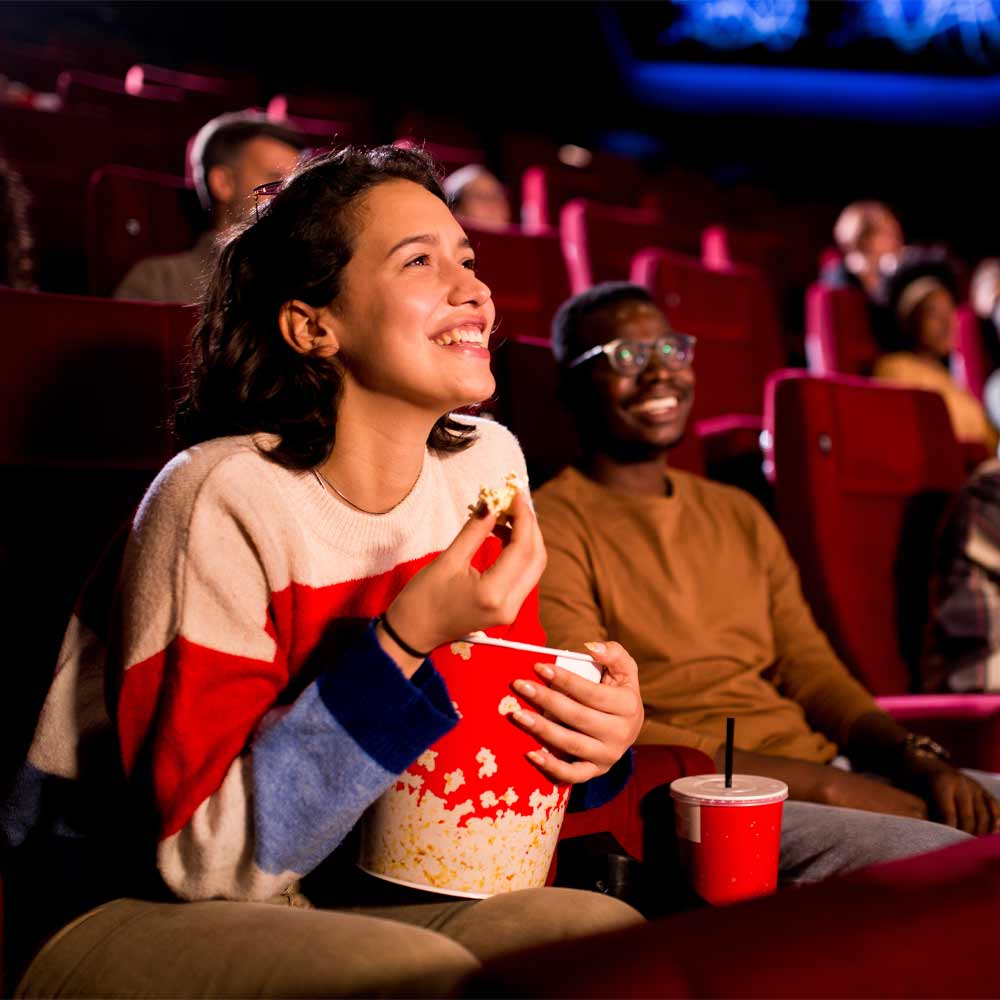 L'image montre un groupe de personnes assises dans un cinéma, appréciant un film avec du pop-corn et des boissons.