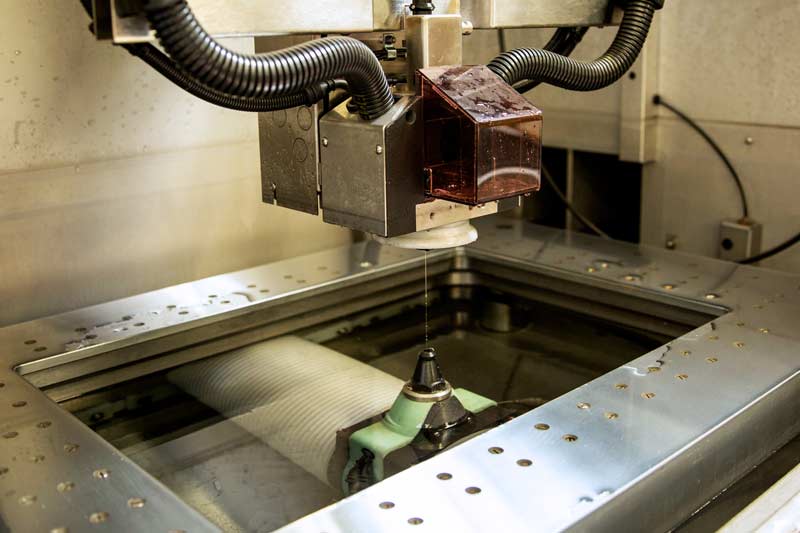 Installation industrielle de découpe par fil électrique (WEDM), montrant la machine, le fil de coupe et la pièce immergée dans le bain, illustrant précision et complexité technique.