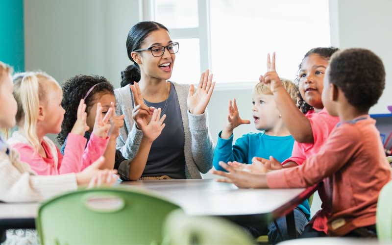 Une enseignante et plusieurs jeunes enfants sont assis autour d'une table et utilisent le langage des signes. L'enseignante, qui porte des lunettes, fait des signes avec ses mains.