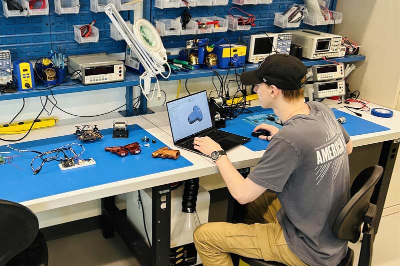 Jeune homme travaillant sur un projet technique dans un atelier moderne, utilisant un ordinateur portable et entouré de composants électroniques et d’outils sur un plan de travail bien équipé.