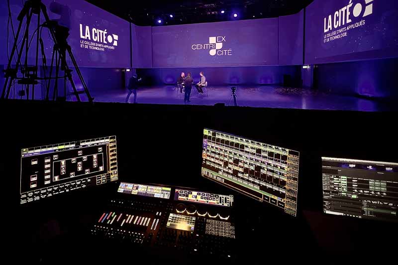 Vue backstage d’un événement ou enregistrement studio, montrant un panneau de contrôle professionnel au premier plan et une scène illuminée avec le texte « LA CITÉ », des participants et du matériel de tournage, illustrant la gestion technique d’une production en direct.