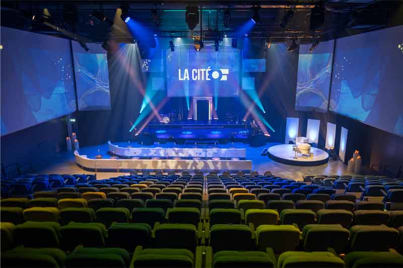 Un auditorium vide prêt pour un événement, avec une scène centrale lumineuse affichant « LA CITÉ », instruments de musique, écrans et éclairages colorés, entouré de sièges disposés symétriquement.