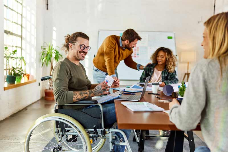 Environnement de travail collaboratif lumineux, avec quatre personnes autour d’une grande table, incluant un homme en fauteuil,   central et souriant, échangeant des documents et utilisant un ordinateur portable dans une ambiance conviviale et inclusive.