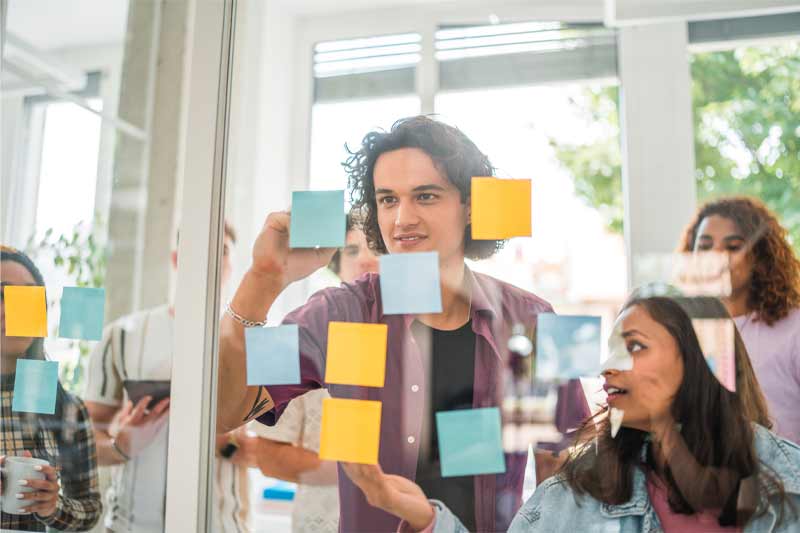 Session de brainstorming collaborative dans un bureau moderne, avec un groupe de personnes échangeant et plaçant des post-it colorés sur une paroi en verre, dans une ambiance lumineuse et interactive.