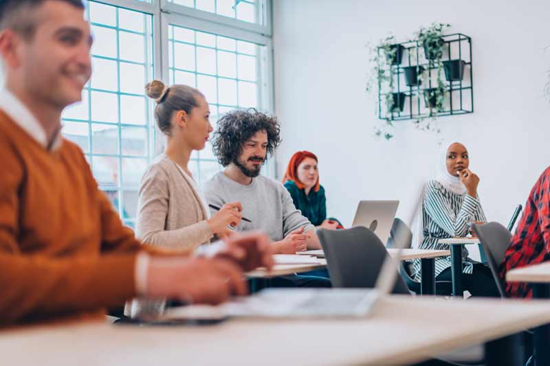 Groupe divers dans une salle lumineuse, collaborant autour de tables avec ordinateurs portables, échangeant activement dans une ambiance positive et participative.