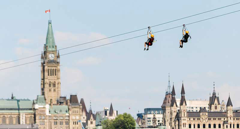 Two people zip line over the Canadian Parliament Buildings, offering a breathtaking view of this historic architecture.