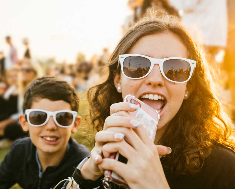 Au premier plan, deux jeunes gens, un garçon et une fille, portant des lunettes de soleil, sont nets. La fille tient son téléphone à la bouche, l'air enthousiaste.