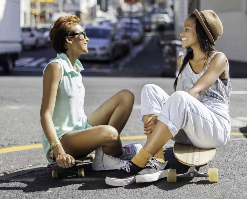 Deux jeunes femmes sont assises sur des skateboards dans la rue, face à face et souriantes. Elles semblent être amies et profitent d'un moment de détente en plein air.
