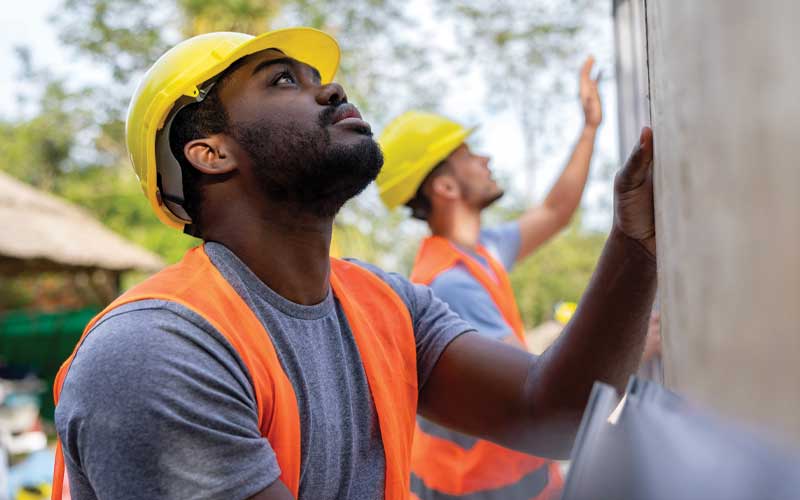 Deux ouvriers du bâtiment, casqués et vêtus de gilets de sécurité, regardent vers le haut. L'un d'eux, plus près de la caméra, touche un mur.