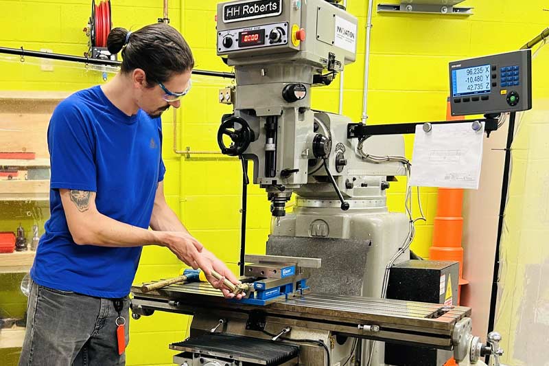 Homme travaillant sur une fraiseuse dans un atelier, portant lunettes de sécurité et concentré sur une pièce métallique, illustrant précision et travail technique.