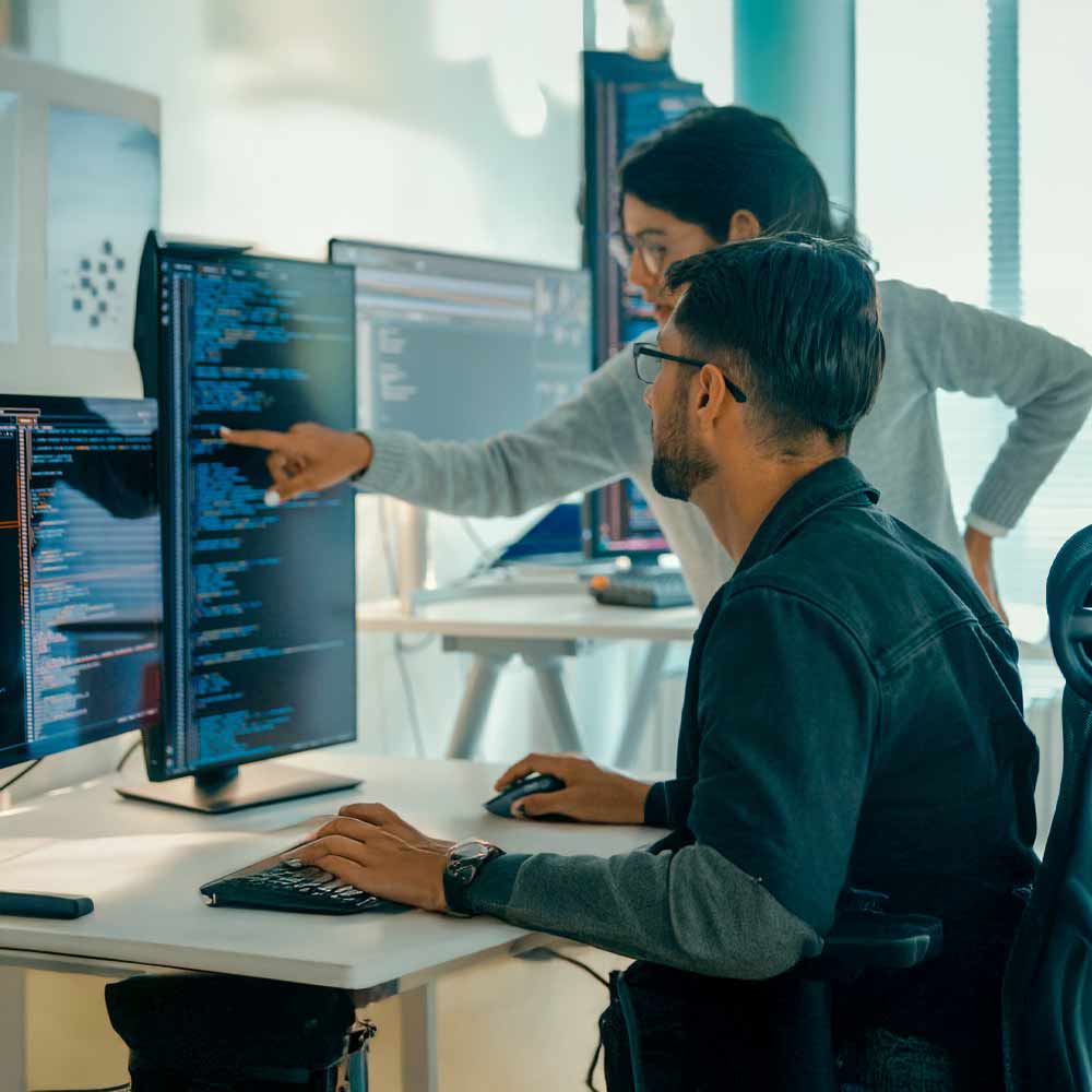 Deux programmeurs collaborant dans un bureau moderne, avec plusieurs écrans affichant du code, l’un assis au clavier et l’autre pointant un écran, illustrant une dynamique de travail concentrée et interactive.