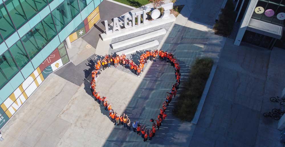 Un groupe de personnes vêtues de t-shirts orange forme un cœur au sol. Derrière elles se trouve un grand panneau en béton sur lequel on peut lire « LA CITÉ ».