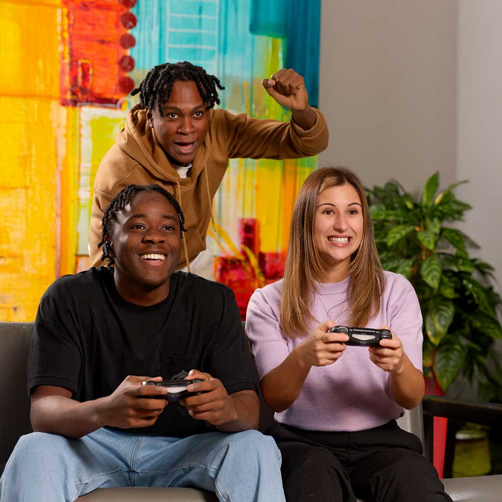 Trois jeunes adultes souriants jouent à un jeu vidéo avec des manettes, assis sur un canapé dans un espace coloré et convivial.