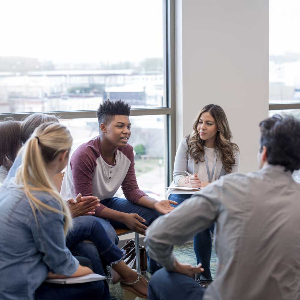 Séance de groupe ou de counseling dans une pièce moderne et lumineuse, centrée sur un cercle de participants attentifs, avec un jeune homme noir prenant la parole au centre et un environnement lumineux et naturel favorisant l’échange.