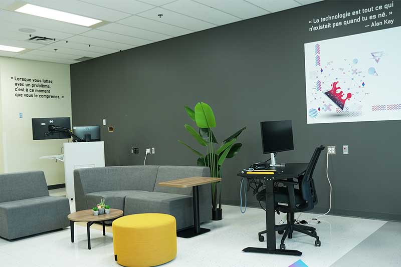 Espace de bureau moderne et lumineux, comprenant un coin détente et un poste de travail. Le coin détente est meublé d'un canapé gris, de chaises, d'une petite table et d'un pouf jaune. Le poste de travail est équipé d'un ordinateur, d'un bureau réglable et d'une chaise noire.