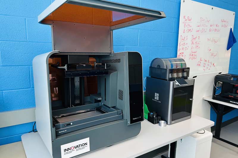 Laboratoire technologique avec une grande imprimante 3D et un équipement supplémentaire sur des tables blanches, dans un espace organisé et moderne dédié à l’innovation.