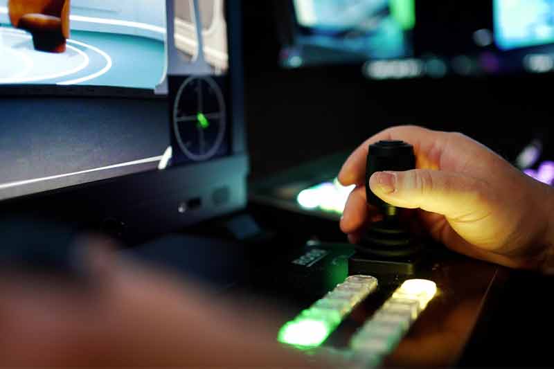 Gros plan sur la main d’un opérateur manipulant un joystick sur une console de contrôle en studio, avec des boutons illuminés et des écrans en arrière-plan, illustrant précision et professionnalisme.