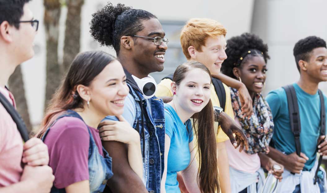 Cinq jeunes amis aux profils variés, sacs à dos sur le dos, sourient joyeusement en posant ensemble à l’extérieur. Bras dessus bras dessous, ils semblent prêts à partir à l’aventure.