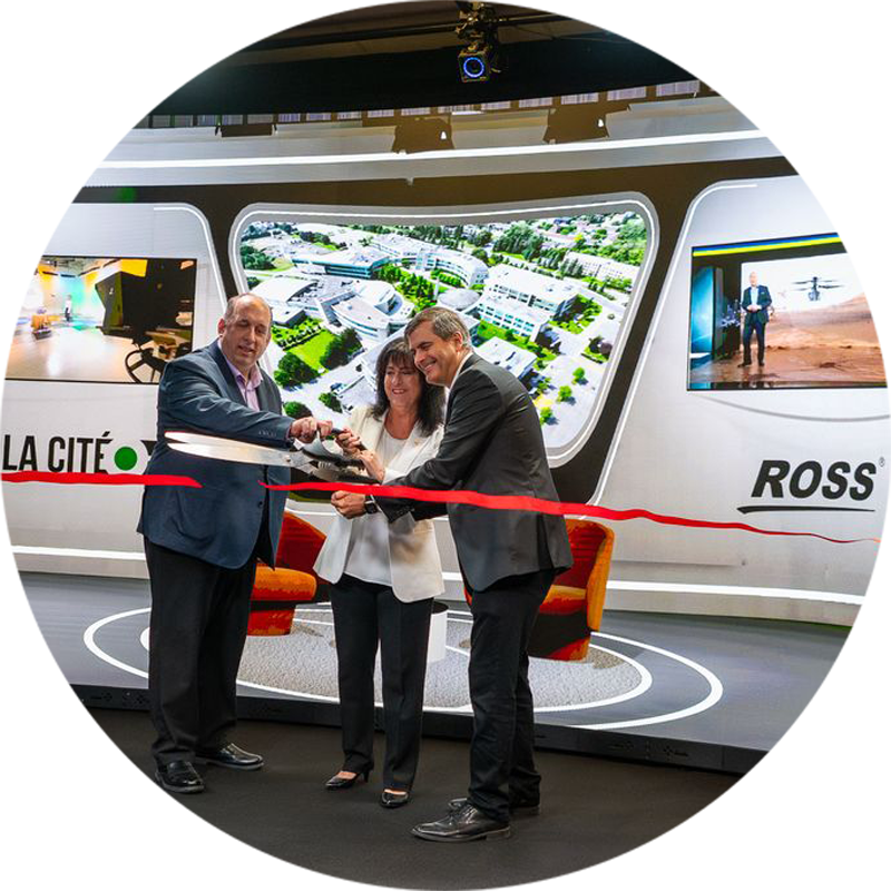 Cérémonie d’inauguration avec trois personnes coupant un ruban rouge devant un écran incurvé affichant « LA CITÉ » et « ROSS », dans un décor moderne et minimaliste comprenant écrans, panneaux blancs incurvés, sièges orange et luminaires au plafond noir.
