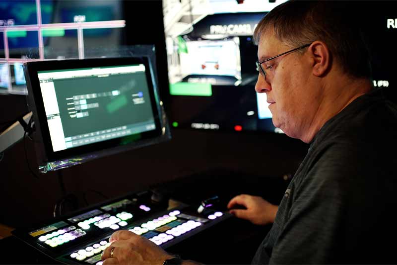 Un homme portant des lunettes manipule un panneau de contrôle doté de plusieurs écrans affichant des informations. Il se trouve dans une salle de contrôle production.