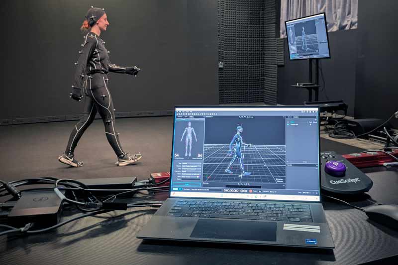 Une femme vêtue d'une combinaison de capture de mouvement marche dans un studio. Un écran d'ordinateur portable affiche un modèle 3D reproduisant ses mouvements, tandis que d'autres moniteurs affichent les données de capture de mouvement.