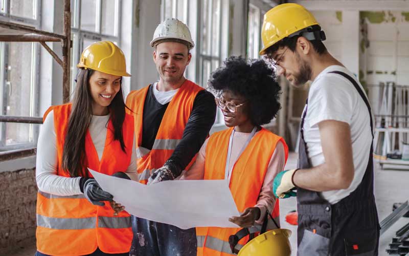 Un groupe hétéroclite d'ouvriers du bâtiment, portant casques et gilets de sécurité, est rassemblé autour d'un plan. Ils semblent en discuter.